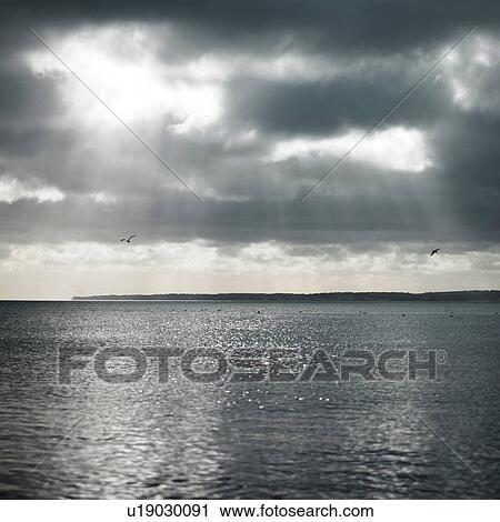 Stock Image - Sunbeams through clouds over ocean. Fotosearch