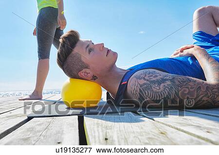 Stock Photo - Tattooed man lying on pier using ball as pillow. Fotosearch