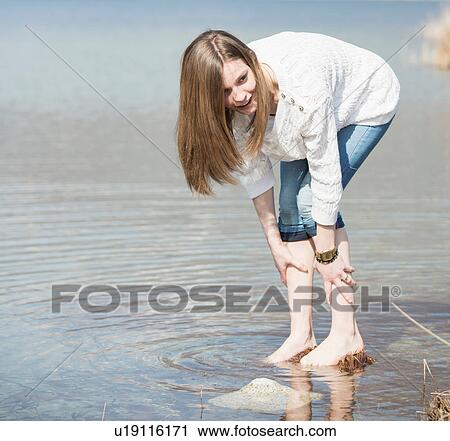 Stock Image - Teenage girl with jeans rolled up ankle deep in water bending forwards. Fotosearch