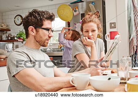Stock Image - Tired mid adult parents at breakfast table whilst daughter playing with balloon. Fotosearch