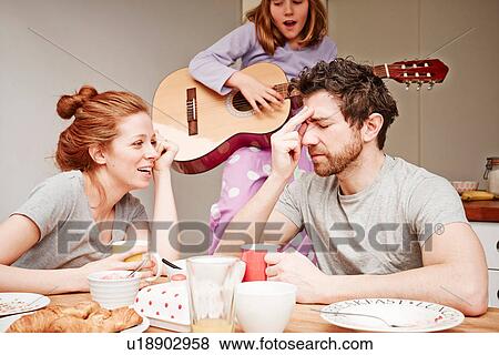 Stock Photo - Tired mid adult parents at breakfast table whilst daughter plays guitar. Fotosearch