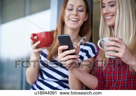 Two female friends, drinking coffee, outdoors, looking at smartphone View Large Photo Image Stock Image - Two female friends, drinking coffee, outdoors, looking at smartphone. Fotosearch