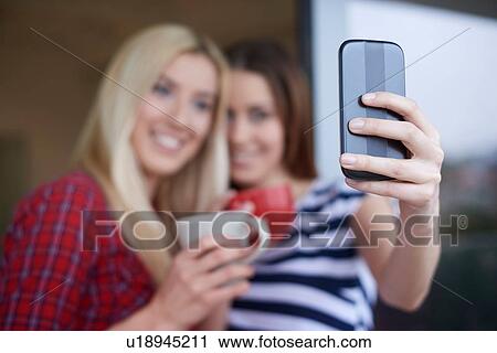 Stock Image - Two female friends, drinking coffee, outdoors, taking self portrait using smartphone. Fotosearch