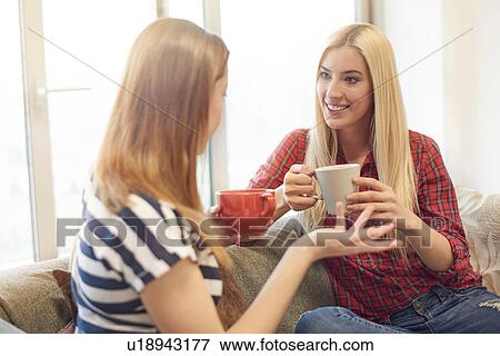 Stock Photo - Two female friends, sitting on sofa, drinking coffee, catching up. Fotosearch