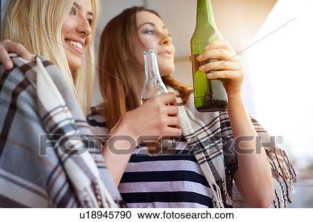 Stock Image - Two female friends, sitting outdoors, drinking bottled beer. Fotosearch