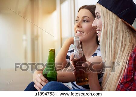 Stock Image - Two female friends, sitting outdoors, drinking bottled beer. Fotosearch