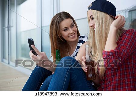 Stock Image - Two female friends, sitting outdoors, drinking bottled beer, looking at smartphone. Fotosearch