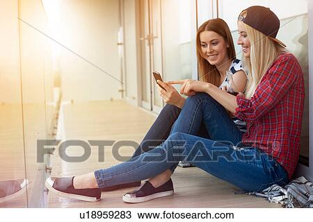 Stock Image - Two female friends, sitting outdoors, looking at smartphone. Fotosearch