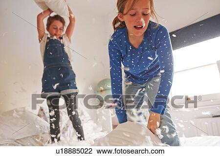 Stock Image - Two sister having feather pillow fight on bed. Fotosearch