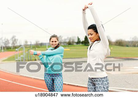 Stock Photo - Two young women on running track, exercising, stretching. Fotosearch