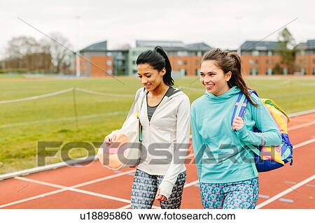 Stock Image - Two young women walking on running track, carrying sports bags. Fotosearch