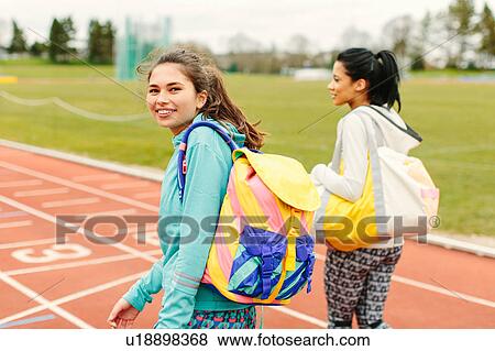Stock Photo - Two young women walking on running track, carrying sports bags, rear view. Fotosearch