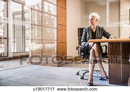 Stock Photo - View through glass of businesswoman sitting at desk legs crossed looking away. Fotosearch