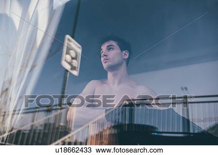 Stock Image - View through glass of man putting on t-shirt, looking away. Fotosearch