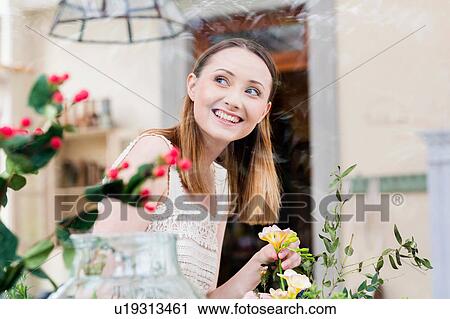 Stock Image - View through glass of woman arranging flowers looking away smiling. Fotosearch