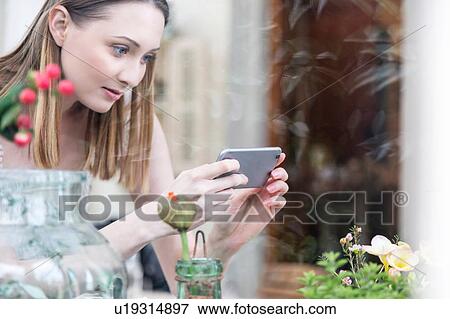 Stock Photo - View through glass of woman using smartphone to photograph plants. Fotosearch