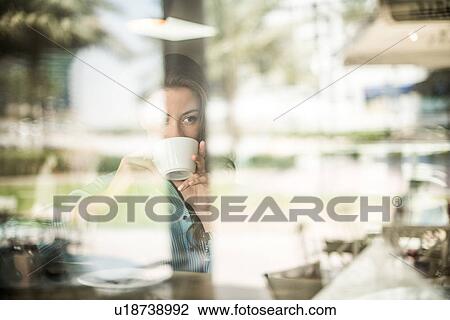 Stock Image - Woman drinking coffee at reflective cafe window, Dubai, United Arab Emirates. Fotosearch