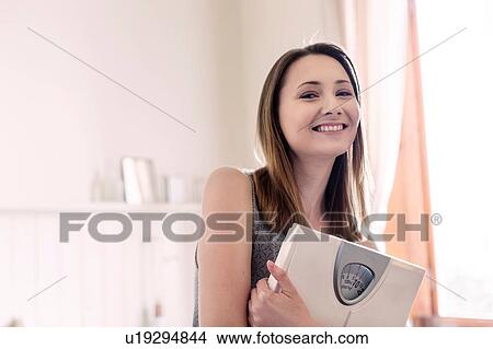 Woman holding bathroom scales looking at camera smiling View Large Photo Image Picture - Woman holding bathroom scales looking at camera smiling. Fotosearch