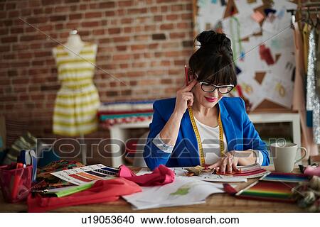 Stock Image - Woman in design studio sitting at desk, hand on head looking down. Fotosearch