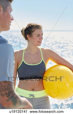 Stock Image - Woman in front of ocean holding yellow ball looking away smiling. Fotosearch