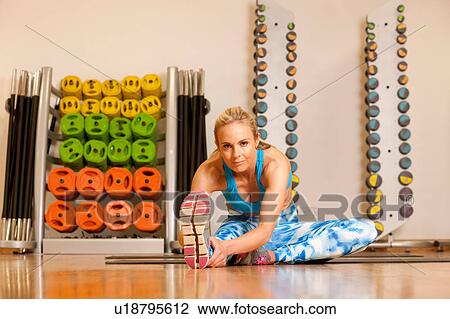 Stock Image - Woman in gym sitting on yoga mat doing leg stretching exercises. Fotosearch