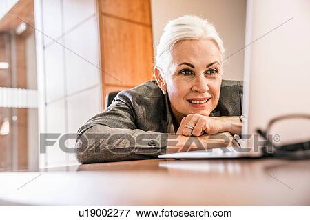 Stock Photo - Woman leaning against desk chin on hands looking at laptop smiling. Fotosearch