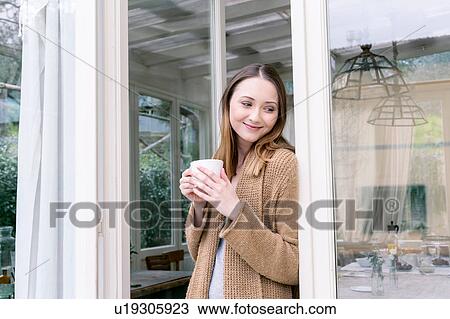 Woman leaning against doorway holding coffee cup looking away smiling View Large Photo Image Stock Image - Woman leaning against doorway holding coffee cup looking away smiling. Fotosearch