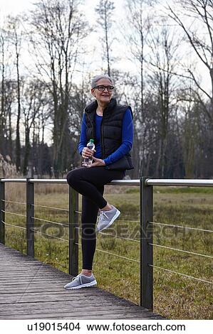 Picture - Woman leaning against railing holding bottle of water looking away. Fotosearch