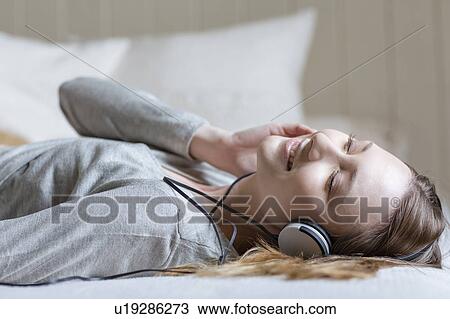 Stock Image - Woman lying on bed wearing head phones, eyes closed smiling. Fotosearch