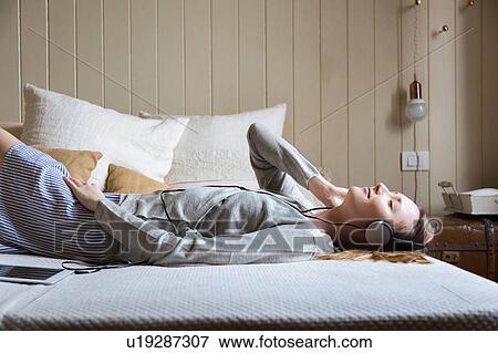 Stock Photo - Woman lying on bed wearing head phones, eyes closed smiling. Fotosearch