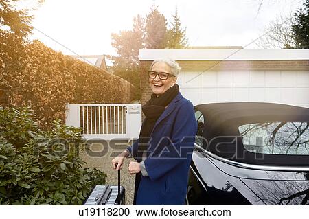 Stock Image - Woman near car on driveway holding suitcase looking at camera smiling. Fotosearch