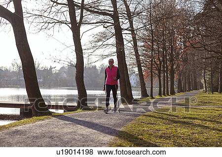 Stock Photo - Woman nordic walking on tree lined path by pond. Fotosearch