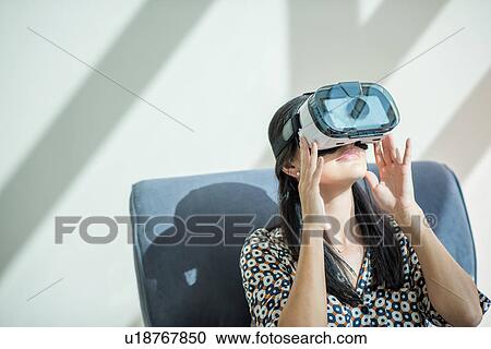 Stock Image - Woman on armchair looking through virtual reality headset. Fotosearch