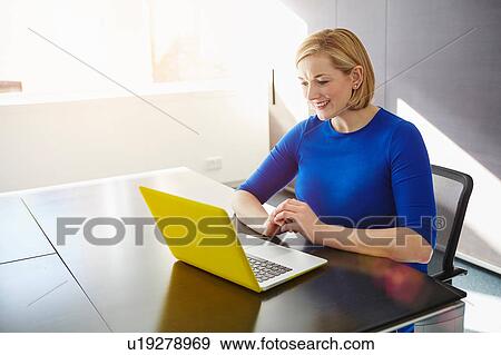 Stock Photo - Woman sitting at desk using laptop smiling. Fotosearch