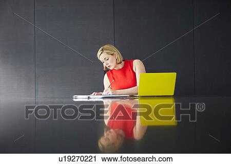 Stock Image - Woman sitting at desk with laptop writing in notebook. Fotosearch