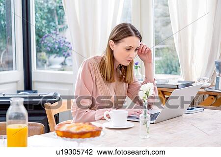 Stock Image - Woman sitting at dining table using laptop. Fotosearch