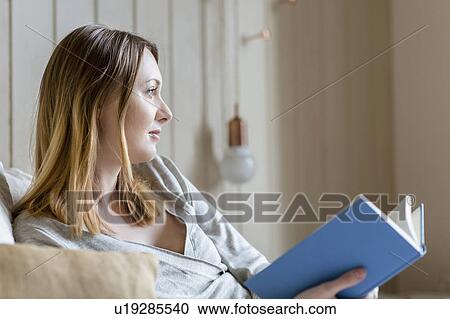 Stock Image - Woman sitting on bed holding book looking away. Fotosearch