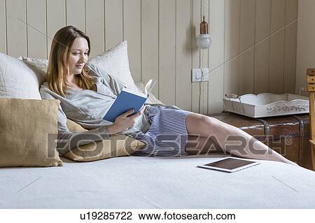 Stock Image - Woman sitting on bed reading book. Fotosearch