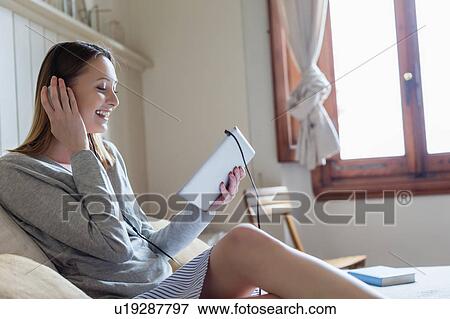 Woman sitting on bed wearing headphone looking at digital tablet smiling View Large Photo Image Stock Photo - Woman sitting on bed wearing headphone looking at digital tablet smiling. Fotosearch