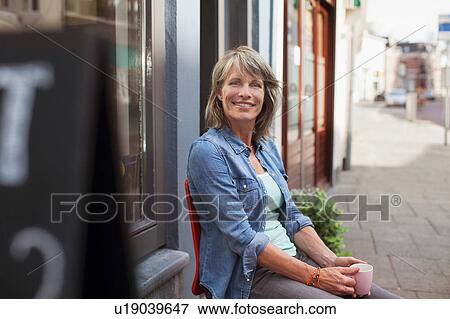 Woman sitting on chair in front of shop holding coffee cup looking at camera smiling View Large Photo Image Stock Photo - Woman sitting on chair in front of shop holding coffee cup looking at camera smiling. Fotosearch