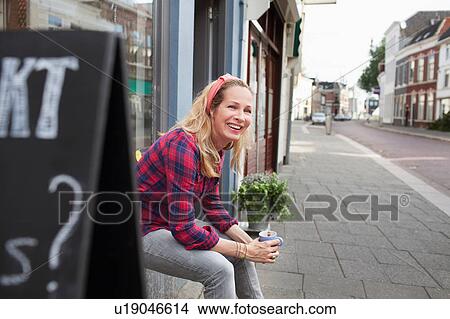Picture - Woman sitting on shop windowsill holding coffee cup looking away smiling. Fotosearch