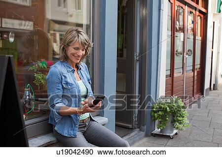 Woman sitting on shop windowsill looking down at smartphone smiling View Large Photo Image Stock Image - Woman sitting on shop windowsill looking down at smartphone smiling. Fotosearch