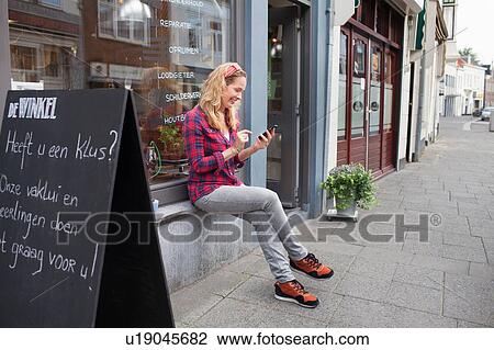 Stock Image - Woman sitting on shop windowsill looking down at smartphone smiling. Fotosearch