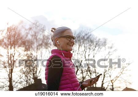 Woman wearing glasses and earbuds, holding smartphone looking away smiling View Large Photo Image Picture - Woman wearing glasses and earbuds, holding smartphone looking away smiling. Fotosearch