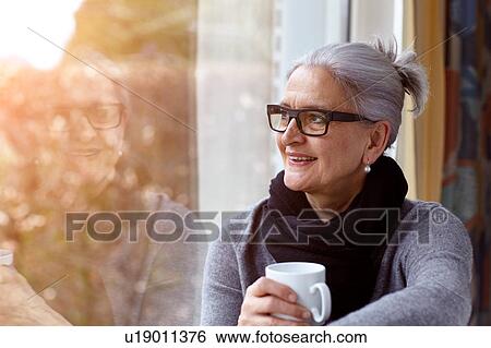 Woman wearing glasses holding coffee cup looking out of window smiling View Large Photo Image Stock Photograph - Woman wearing glasses holding coffee cup looking out of window smiling. Fotosearch