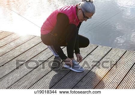 Picture - Woman wearing pink bodywarmer and earbuds crouching on pier tying shoelace. Fotosearch