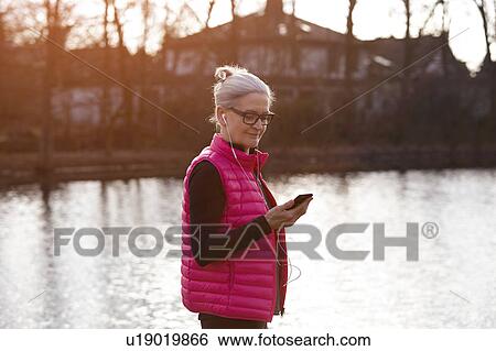 Stock Photograph - Woman wearing pink bodywarmer and earbuds in front of pond looking at smartphone. Fotosearch
