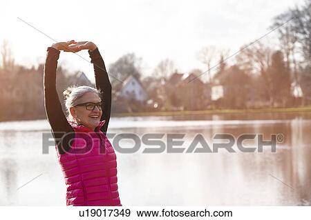 Stock Photo - Woman wearing pink bodywarmer in front of pond arms raised stretching looking away smiling. Fotosearch