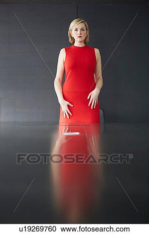 Stock Image - Woman wearing red dress standing at desk looking at camera. Fotosearch