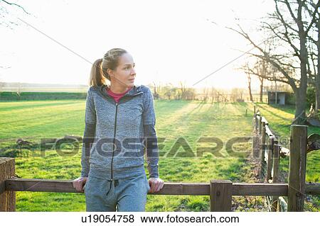 Stock Photo - Woman wearing sports clothing leaning against fence looking away. Fotosearch